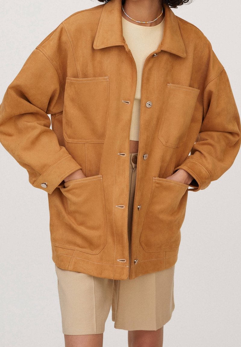 Femme portant une veste en daim ample couleur camel avec de grandes poches avant sur un crop top beige et un short assorti, mains dans les poches.