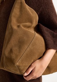 Hnědá semišová kabelka s měkkým, strukturovaným povrchem, mírně sklápějícím tvarem a minimalistickým designem. Obsahuje jemný detail švu.