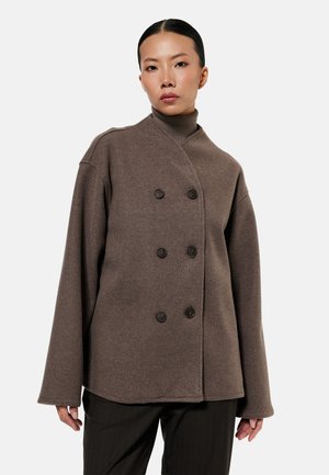 Frau trägt braunen doppelt geknöpften Wollmantel über braunem Rollkragenpullover, kombiniert mit schwarzen Nadelstreifenhosen, vor schlichtem weißem Hintergrund.