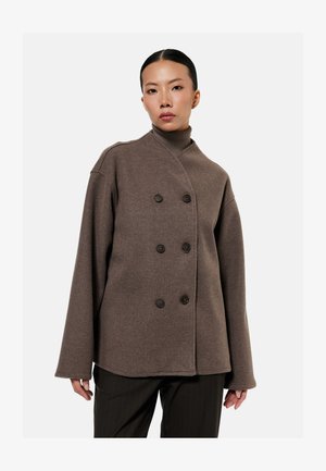 Frau trägt braunen doppelt geknöpften Wollmantel über braunem Rollkragenpullover, kombiniert mit schwarzen Nadelstreifenhosen, vor schlichtem weißem Hintergrund.