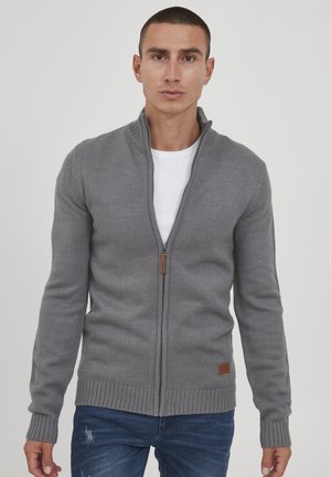 Grauer Reißverschluss-Pullover mit gerippten Bündchen und Saum, ausgestattet mit einem hohen Kragen und einem braunen Lederakzent in der Taille. Wird über einem weißen Hemd getragen.