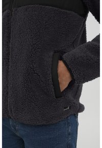 Mørkegrå fleecejakke med glidelås foran, sorte detaljer og sidelommer. Stoffet er teksturert og mykt å ta på.