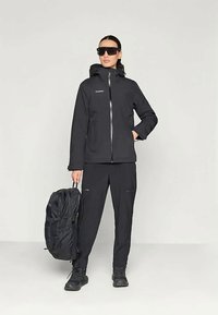 Mulher com uma jaqueta capuz preta, calças pretas, botas e óculos de sol, segurando uma mochila preta ao seu lado, contra um fundo simples.