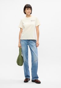 Κρέμα βαμβακερή μπλούζα κοντομάνικη με γραφικό τύπωμα, συνδυασμένη με ανοιχτό μπλε παντελόνι σε φαρδιά γραμμή. Κρατάει μια πράσινη τσάντα με κεντήματα.