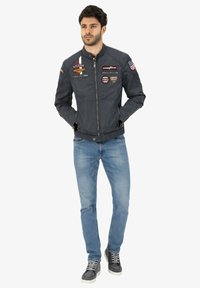 Navy bomber jacket with patches, zip closure, standing collar, and side pockets. Paired with light blue jeans and gray high-top sneakers.