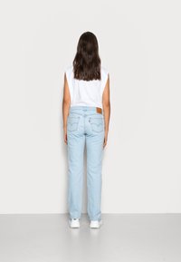 Jeans de ganga de cor azul claro com corte reto, apresentando bolsos traseiros e uma etiqueta de marca em pele castanha. Top sem mangas branco visível.