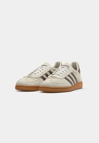 Sneaker beige in suede con tre strisce marroni scure, lacci bianchi e suola in gomma naturale, caratterizzati da una punta arrotondata e una finitura testurizzata.