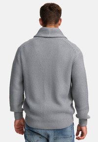 Gestrickter grauer Pullover mit hohem Kragen, gerippten Bündchen und Saum sowie einer strukturierten Oberfläche. Die Rückseite zeigt ein markantes gewebtes Muster.