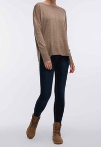 Beige gebreide trui met een losse pasvorm, geribbelde manchetten en zijsplitten, gecombineerd met donkerblauwe skinny jeans en beige veterschoenen voor de enkel.