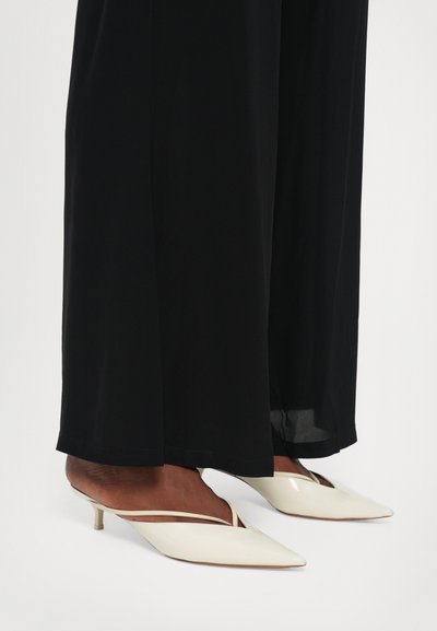 Mules bianchi con punta a stivaletto e tacco kitten sottile, caratterizzati da un design con ritagli e una finitura lucida, abbinati a pantaloni neri fluidi.