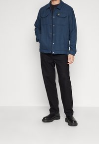 Giacca a scacchi blu con chiusura a bottone e due tasche sul petto, abbinata a pantaloni neri e stivali neri robusti. Dettaglio del tessuto texture evidente.