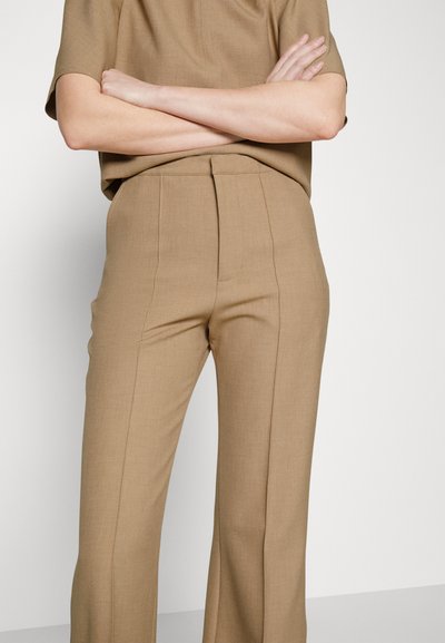 Inimesel on käed risti, seljas beezid kohandatud püksid ja sobiv lühikeste varrukatega särk, taustal üksikasjalik taust.