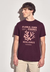 Vínová tričko s korálovým grafickým designem a textem "FLORA A FAUNA" a "SEAL COVE KALIFORNIE". Krátké rukávy, kulatý výstřih a bavlněný materiál.