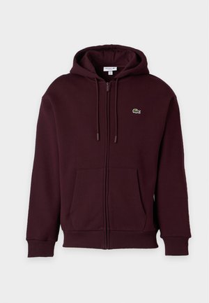Homme debout portant un sweat à capuche bordeaux et un t-shirt, un pantalon gris et des baskets blanches sur un fond blanc uni.