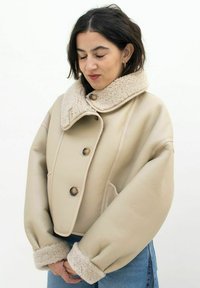 Beige leren jack met een fluffy beige kraag en manchetten, voorzien van twee knopen en een gestructureerd, oversized silhouet.