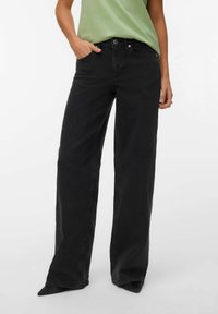 Personne portant un jean noir taille haute à jambes larges, une main dans la poche, associé à un haut vert clair rentré et des chaussures noires à bouts pointus.