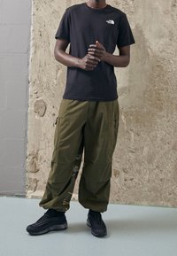 Persona vistiendo una camiseta negra de The North Face, pantalones cargo verde oliva con diseño bordado y zapatillas negras, de pie en el interior sobre un suelo claro.