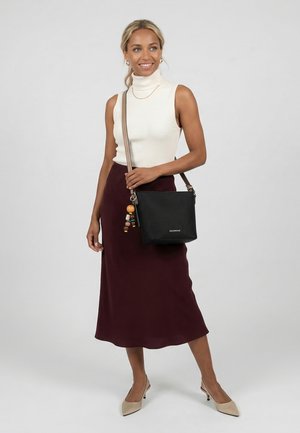 Žena v bezrukávovém bílém roláku, tmavě fialské sukni, béžových lodičkách, nesoucí černou texturovanou kabelku přes rameno s dřevěným přívěskem na klíče.