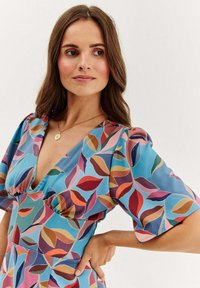 Modré květinové šaty s hlubokým výstřihem do V, krátkými nabíranými rukávy a vzorem vícebarevných listů v růžových, červených, zelených a hnědých odstínech.