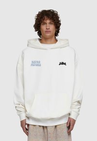 Vit oversized hoodie med en framlapp; pryds av blå "RETRO FUTURE" och svarta skrivställningar. Slät textur med en stor huva.