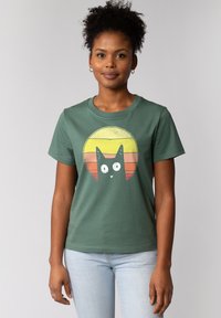 Groene katoenen t-shirt met een afbeelding van een zwarte kat binnen een meerkleurige cirkel met gele en oranje horizontale strepen. Korte mouwen.