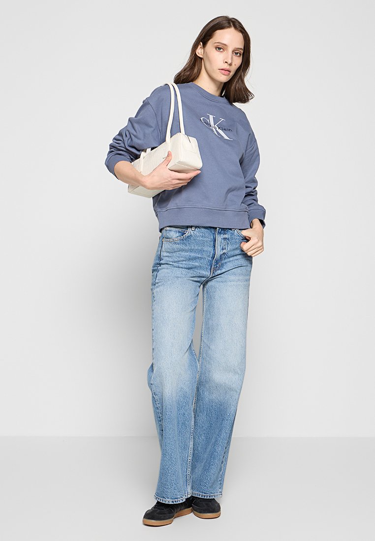 Felpa blu con logo sul petto, abbinata a jeans chiari a zampa d'elefante. Una piccola borsa bianca e scarpe nere completano il look.