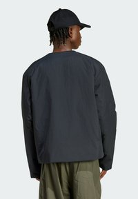 Osoba s krútenými vlasmi, ktorá má na sebe čiernu čiapku, čiernu dlhú bundu a olivové zelené nohavice, stojí chrbtom k jednoduchému pozadiu.