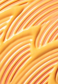 Prodotto cremoso di colore pesca applicato con colpi morbidi e ondulati su una superficie rosa chiaro, caratterizzato da una texture lucida.