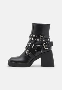 Bottines noires à talon épais, ornées de clous en argent et de boucles sur les sangles. Fini cuir lisse avec une silhouette épurée.