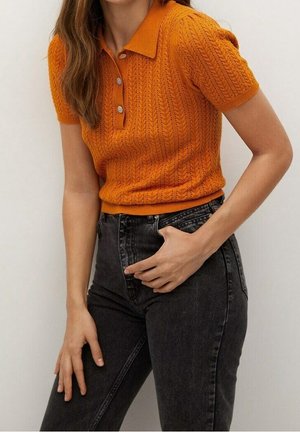 Kvinde iført en kortærmet, orange strikket poloskjorte med knapper og sort højtaljede jeans, poserer med den ene hånd i lommen.