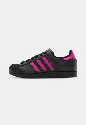 Baskets en cuir noir avec trois rayures roses, embout en caoutchouc et semelle plate noire. Présente un design à lacets et le texte "SUPERSTAR" sur le côté.