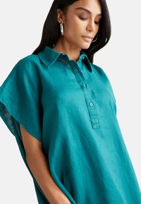 Turkusowa lniana koszula z krótkimi rękawami, z zapięciem na guziki i teksturowanym materiałem. Charakteryzuje się luźnym krojem i widocznymi detalami szwów.