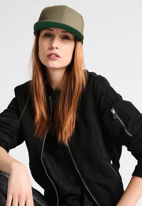 Zwarte bomberjack met ritssluitingen, gecombineerd met een groene snapback pet. Model heeft lang, steil haar. Donkere achtergrond.
