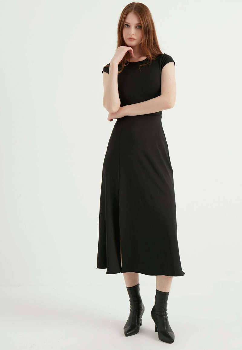 Schwarzes, knielanges Kleid mit kurzen Ärmeln, einem taillierten Oberteil und einem asymmetrischen Schlitz vorne. Glattes Material und klares Design.