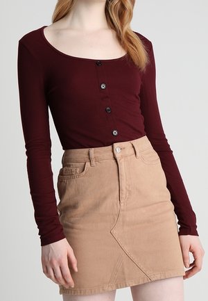 Kobieta w dopasowanej bordo bluzce z długim rękawem z guzikami oraz w beżowej spódnicy z wysokim stanem, z przeszyciami z przodu i kieszeniami.
