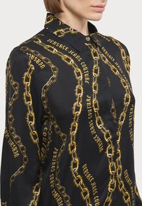 Camisa preta com um padrão de corrente dourada e texto, mangas longas, colarinho pontiagudo e fecho de botões com acabamentos metálicos.