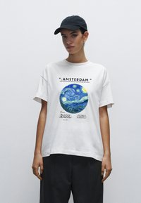 Bílé bavlněné tričko s modro-žlutou kruhovou grafikou se vzorem hvězdné noci a textem o Amsterdamu, nošené s šedými volnými kalhotami.