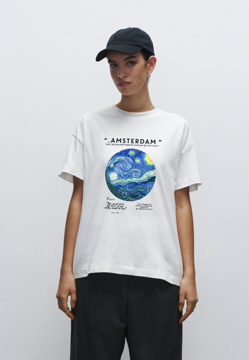 Bílé bavlněné tričko s modro-žlutou kruhovou grafikou se vzorem hvězdné noci a textem o Amsterdamu, nošené s šedými volnými kalhotami.