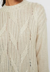 Pull beige en maille torsadée avec col rond côtelé, présentant un motif texturé et des manches longues.