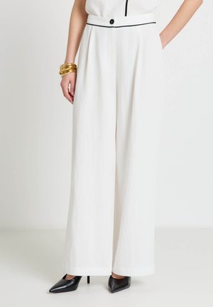 Woman wearing high-waisted, wide-leg white trousers with black piping, black button and black high-heeled shoes, accessorized with gold bangles.