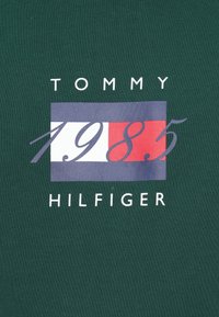 Tamsiai žalias audinio marškinėlių tipo T-shirt su apvaliu kaklu, kuriame vaizduojamas grafikas su "TOMMY HILFIGER" ir "1985" balta ir raudona spalvomis ant mėlyno fono.