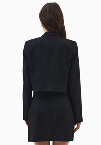 Blazer nero con righe sottili e design corto. Presenta maniche lunghe e una vestibilità sartoriale. Include un dettaglio con cintura in vita.
