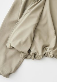 Giacca beige con cerniera argentata parzialmente aperta, che mostra una fodera interna liscia e un orlo inferiore arricciato ed elasticizzato.