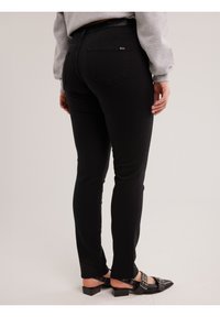 Persona vistiendo jeans negros de corte ajustado, sudadera gris y zapatos negros de tacón bajo con hebillas, de pie contra un fondo claro y liso.