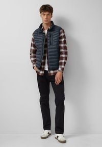 Dunkelblauer Herren-Puffervest, über einem rot-weiß karierten Hemd getragen. Enganliegende schwarze Hose und weiße Sneakers mit grauen Akzenten runden das Outfit ab.