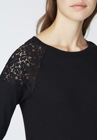 Musta neulepaita, jossa pitsiyksityiskohtia olkapäillä, pehmeä neulekudos ja klassinen pyöreä pääntie. Lähikuva korostaa mallia.