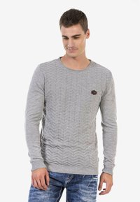 Grauer Pullover mit strukturiertem Chevron-Muster, Rundhalsausschnitt und langen Ärmeln. Besitzt ein kleines, sechseckiges Logo auf der Brust. Getragen mit blauen Jeans.
