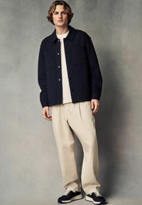 Marinefarbener strukturierter Blazer mit zwei frontalen Taschen, weißem Rundhalsausschnitt-T-Shirt, beigen locker sitzenden Hosen und marineblauen Sneakern mit weißen Schnürsenkeln.