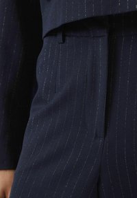 Marynarskie spodnie z teksturowanej tkaniny, z subtelnymi prążkami i metalicznymi akcentami, wyeksponowane obok dopasowanej marynarki.
