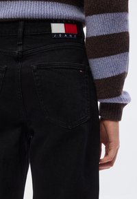 Μαύρο τζιν από τζιν με ψηλή μέση, με πίσω τσέπες και ετικέτα με λογότυπο σε κόκκινο, άσπρο και μπλε στη ζώνη. Φανερό ριγέ πουλόβερ.